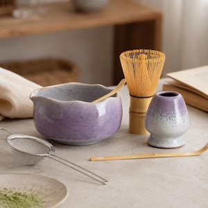 May include: A matcha tea set featuring a lavender ceramic bowl with a scalloped rim, a bamboo whisk, a matching lavender whisk holder, a bamboo scoop, and a stainless steel mesh strainer. A small ceramic plate with green matcha powder is included.
