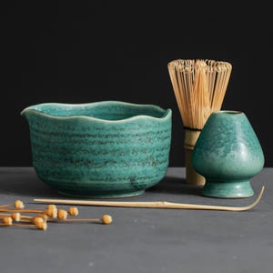 May include: A teal ceramic matcha bowl with a wavy rim, a bamboo whisk, a matcha scoop, and a small teal ceramic matcha container. The items are arranged on a gray surface against a dark background. Dried seed pods are in the foreground.