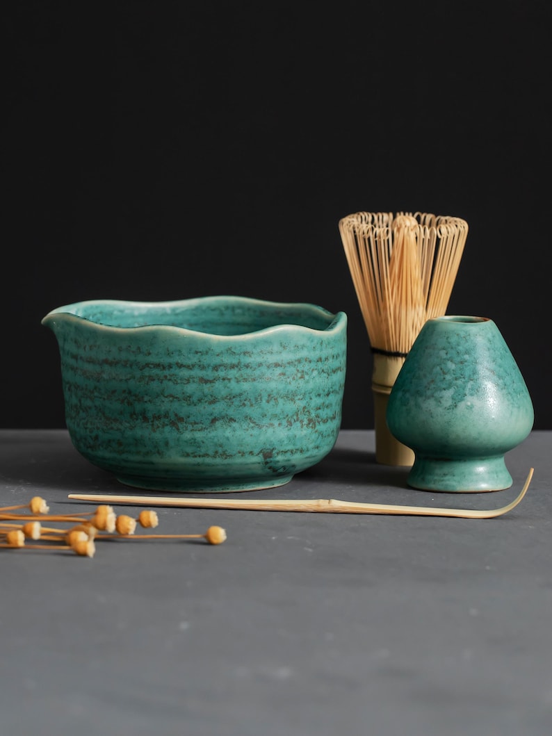 May include: A teal ceramic matcha bowl with a wavy rim, a bamboo whisk, a matcha scoop, and a small teal ceramic matcha container. The items are arranged on a gray surface against a dark background. Dried seed pods are in the foreground.