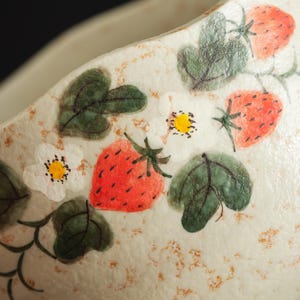 May include: A cream-colored ceramic bowl featuring a hand-painted pattern of red strawberries, green leaves, and white flowers with yellow centers. The surface has a textured, speckled appearance with light brown flecks across the exterior.