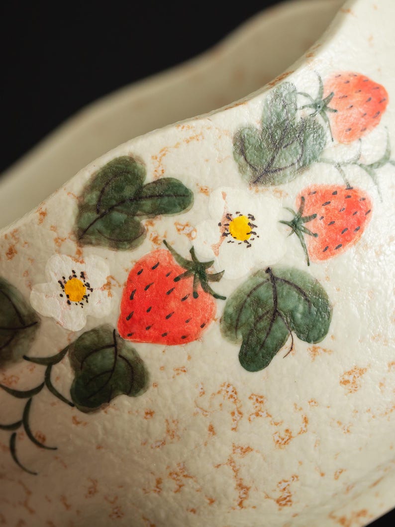 May include: A cream-colored ceramic bowl featuring a hand-painted pattern of red strawberries, green leaves, and white flowers with yellow centers. The surface has a textured, speckled appearance with light brown flecks across the exterior.