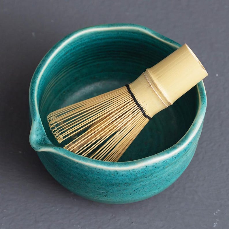 May include: A teal ceramic matcha bowl with a bamboo whisk. The bowl has a slightly irregular shape and a small spout. The whisk is made of thin bamboo tines bound together with black thread. The background is a solid gray.