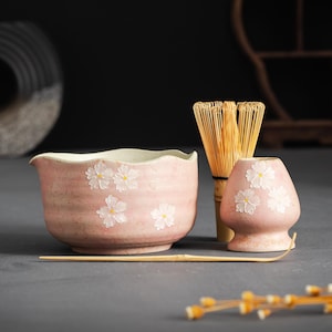 Op de afbeelding: Een roze keramische matcha kom en accessoires set. De kom en kleine container hebben witte bloemmotieven. Een bamboe klopper en lepel zijn ook inbegrepen, alles tegen een donkere achtergrond.