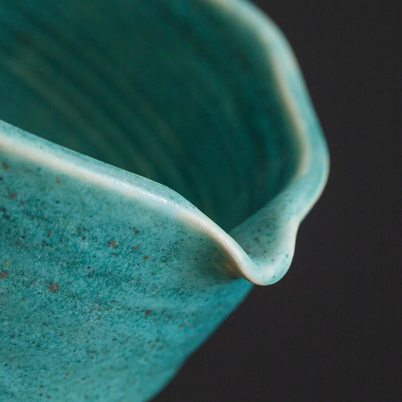 May include: Close-up of a teal ceramic bowl with a textured surface and a white rim. The bowl has a curved, organic shape and a speckled appearance, suggesting a handmade quality. The background is dark.