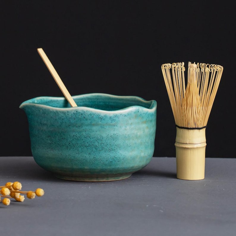 May include: A teal ceramic matcha bowl with a wavy rim and a wooden spoon. A bamboo matcha whisk stands beside the bowl. The items are on a gray surface with a dark background. The bowl is approximately 4 inches in diameter.