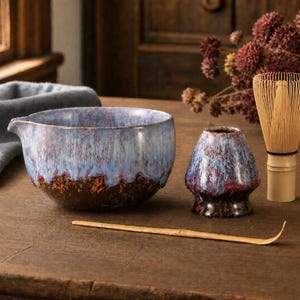 Könnte beinhalten: Eine Matcha-Schale aus Keramik mit blau-brauner Tropfglasur, ein passender Besenhalter, ein heller Bambusbesen und ein dünner Bambuslöffel. Die Schale hat einen kleinen Ausguss an einer Seite und das Set zeigt eine gesprenkelte Oberfläche.