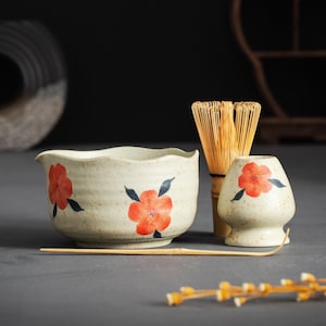 Op de afbeelding: Een matcha-theeset met een keramische kom, een klopper en een kleine houder. De crèmekleurige keramische stukken zijn versierd met rode bloemmotieven en blauwe accenten. Een bamboe klopper en lepel zijn ook inbegrepen.
