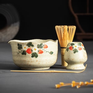 May include: A ceramic matcha bowl with a wavy rim and a matching whisk holder, both featuring a hand-painted strawberry pattern on a speckled cream background. A bamboo whisk and a thin, curved bamboo tea scoop are included in the set.
