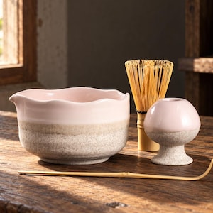 Op de afbeelding: Een keramische matchakom met een golvende rand, voorzien van een glad lichtroze bovendeel en een gestructureerde beige gespikkelde basis. Inclusief een bamboe garde, een bijpassende roze en beige gardehouder en een gebogen bamboe theeschep.