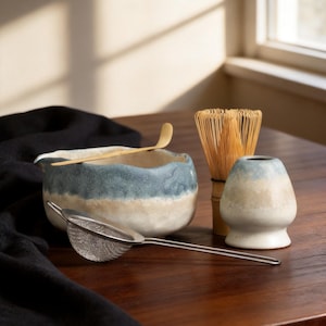 Puede incluir: Un cuenco de cerámica para matcha con esmalte degradado en azul y crema, un batidor de bambú, una cuchara de bambú, un soporte de cerámica para el batidor en azul y crema, y un colador de malla fina de acero inoxidable con mango largo.