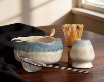 Puede incluir: Un cuenco de cerámica para matcha con esmalte degradado en azul y crema, un batidor de bambú, una cuchara de bambú, un soporte de cerámica para el batidor en azul y crema, y un colador de malla fina de acero inoxidable con mango largo.
