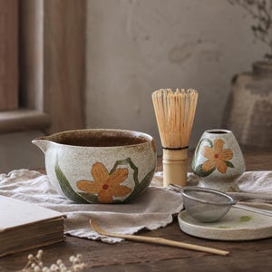 Könnte beinhalten: Eine Matcha-Schale aus Keramik mit gesprenkeltem cremefarbenem Finish und orangefarbenen Blumenmotiven, ein Bambusbesen, ein passender Besenhalter, ein kleines Metallsieb auf einem Keramikteller und ein langer Bambuslöffel für Matcha.