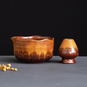 May include: A ceramic matcha bowl with a wavy rim and a matching whisk holder, both featuring a gradient glaze that transitions from a warm golden yellow at the top to a deep, dark brown at the base with a glossy, mottled finish throughout.
