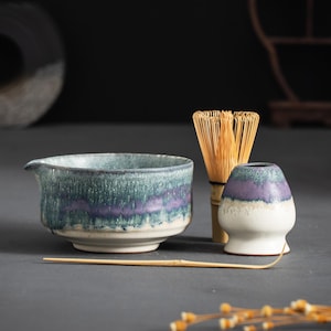 Op de afbeelding: Een keramische matcha kom met een blauw en paars glazuur, een bamboe klopper, een matcha lepel en een keramische matcha kom rust. De theeset staat op een grijze ondergrond.