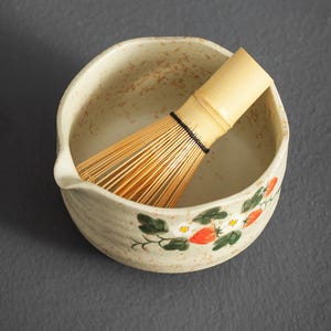 May include: A beige ceramic matcha bowl with a speckled finish and a hand-painted strawberry and white flower motif, featuring a small pouring spout. Inside the bowl rests a light-colored bamboo matcha whisk with thin, flexible tines.