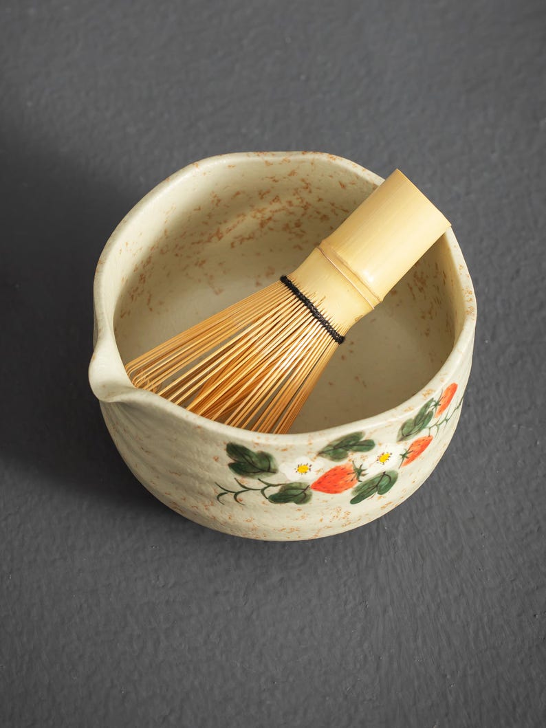 May include: A beige ceramic matcha bowl with a speckled finish and a hand-painted strawberry and white flower motif, featuring a small pouring spout. Inside the bowl rests a light-colored bamboo matcha whisk with thin, flexible tines.