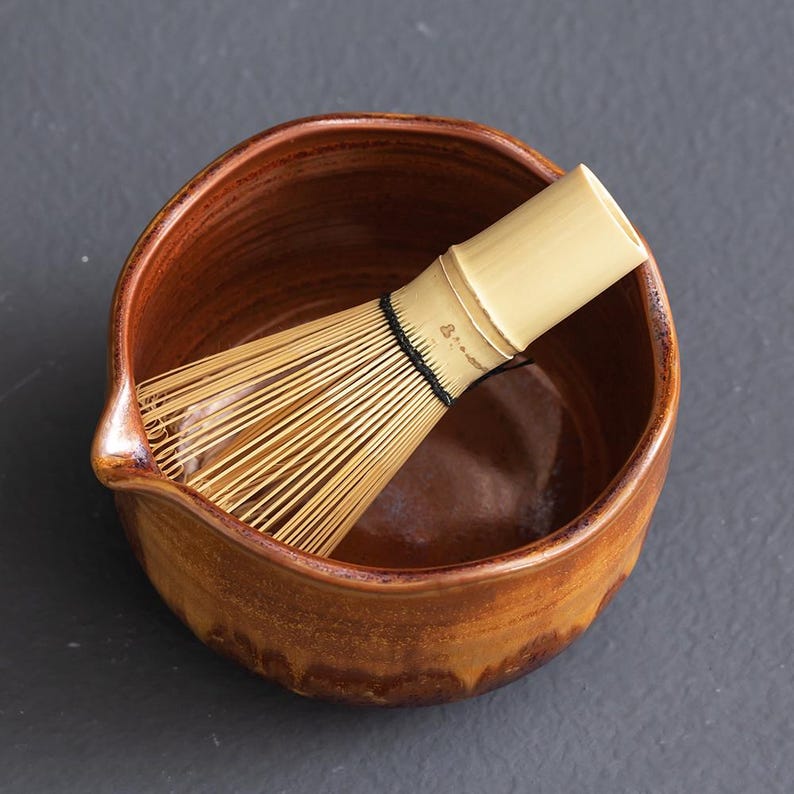 May include: A brown ceramic bowl with a spout and a light-colored bamboo whisk featuring thin, flexible tines tied with a black thread. The bowl has a smooth, glazed interior and a slightly textured, earthy exterior finish, holding the whisk inside.