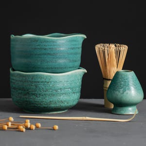 May include: Two teal ceramic bowls with wavy rims are stacked. A bamboo whisk, a small teal ceramic container, and a bamboo spoon are next to the bowls. Dried flower buds are scattered in the foreground. The items are on a gray surface.