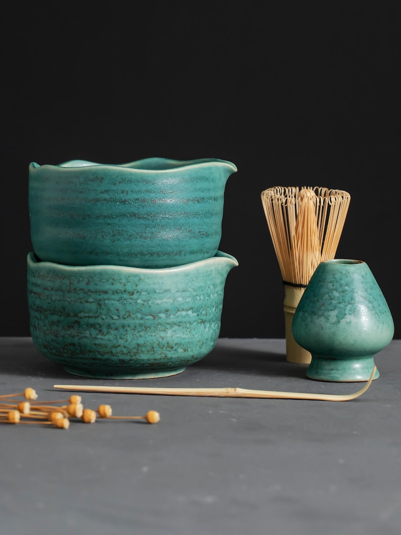 May include: Two teal ceramic bowls with wavy rims are stacked. A bamboo whisk, a small teal ceramic container, and a bamboo spoon are next to the bowls. Dried flower buds are scattered in the foreground. The items are on a gray surface.