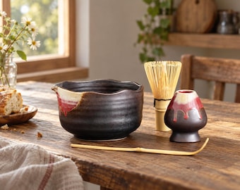 Könnte beinhalten: Eine dunkelbraune Matcha-Schale aus Keramik mit gewelltem Rand und einer cremefarbenen sowie roten Tropfglasur, begleitet von einem Bambusbesen, einem passenden Halter und einem dünnen Bambuslöffel, alles auf einer Holzoberfläche angeordnet.