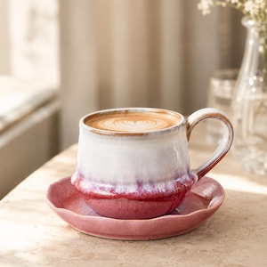 May include: A ceramic mug filled with latte art sits on a matching saucer. The mug features a white upper section that transitions into a deep red base with a glossy, dripped glaze effect. The saucer has a scalloped edge and a solid pink finish.