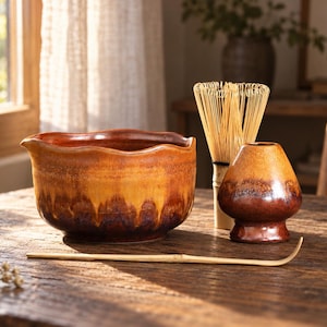 Puede incluir: Un cuenco de cerámica para matcha con borde ondulado y un degradado de esmalte en tonos ámbar, naranja y marrón oscuro. Junto a él, un batidor de bambú, un soporte de cerámica a juego y una cuchara de té de bambú de color claro.