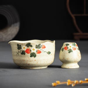 May include: A cream-colored ceramic tea bowl with a spout and a matching whisk holder, both featuring a hand-painted pattern of red strawberries, green leaves, and small white flowers with yellow centers, set against a speckled, textured surface.
