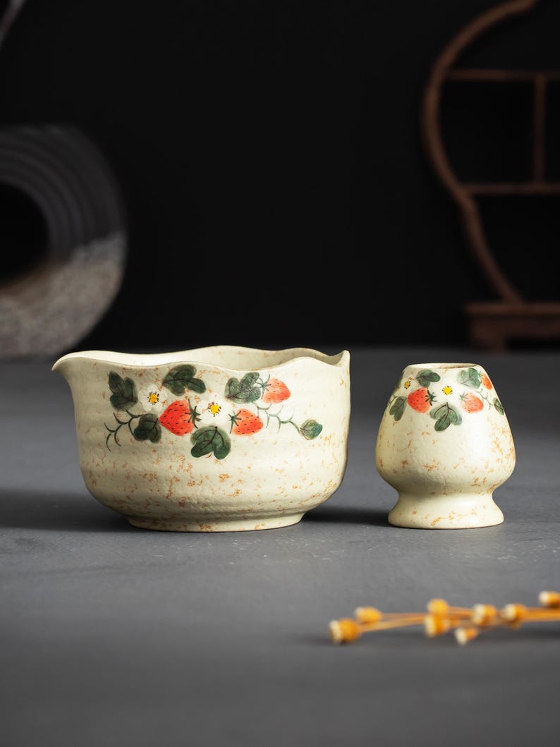 May include: A cream-colored ceramic tea bowl with a spout and a matching whisk holder, both featuring a hand-painted pattern of red strawberries, green leaves, and small white flowers with yellow centers, set against a speckled, textured surface.