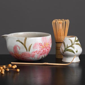 Puede incluir: Un cuenco de té de cerámica blanca con un diseño floral con lirios rosas. El cuenco está al lado de un batidor de bambú y una taza de té de cerámica blanca con un diseño de hoja verde.