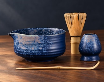 Puede incluir: Un cuenco de matcha de cerámica azul con acabado moteado y pico vertedor, un batidor de bambú, un soporte para batidor de cerámica azul a juego y una cuchara de té de bambú larga, fina y curva.