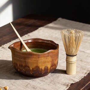 May include: A brown ceramic bowl with a wavy rim containing green matcha tea, a light-colored wooden scoop resting inside, and a bamboo whisk with thin, flexible tines standing upright next to the bowl on a textured fabric surface.