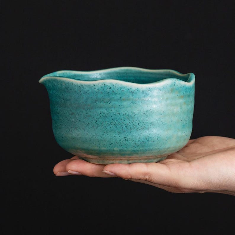 May include: A handmade, turquoise ceramic bowl with a wavy rim and a small spout. The bowl has a textured surface and is held in a hand against a black background. The bowl is likely used for serving or as a decorative piece.