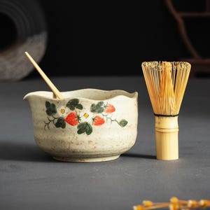 May include: A beige ceramic matcha bowl with a wavy rim, decorated with painted red strawberries and green leaves, accompanied by a light-colored bamboo whisk and a thin wooden stirring stick resting inside the bowl.