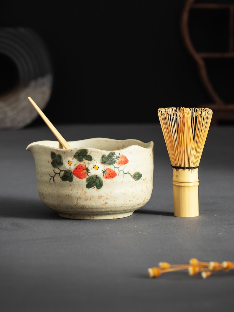 May include: A beige ceramic matcha bowl with a wavy rim, decorated with painted red strawberries and green leaves, accompanied by a light-colored bamboo whisk and a thin wooden stirring stick resting inside the bowl.