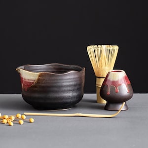 Puede incluir: Un cuenco de matcha de cerámica negro y rojo con un batidor de bambú y una pequeña taza de té de cerámica. También se incluye una cuchara de matcha de madera.