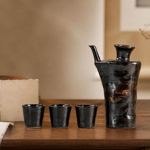 Puede incluir: Un juego de sake de cerámica oscura y texturizada que consta de una jarra alta asimétrica con pico lateral y tres tazas cilíndricas pequeñas a juego. Las piezas presentan un acabado brillante y moteado en tonos negro y marrón.