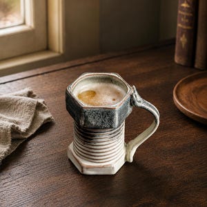 May include: A ceramic mug shaped like a large metal bolt with a hexagonal top rim and a threaded cylindrical body. The mug features a dark, mottled grey glaze on the upper section and a lighter, off-white finish on the lower threaded base.