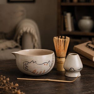 Puede incluir: Un cuenco de cerámica para matcha con una ilustración pintada a mano de un gato naranja y negro, un batidor de bambú, un soporte de cerámica para el batidor con un pájaro y una cuchara de té de bambú. El conjunto tiene un acabado beige moteado.