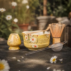 May include: A yellow ceramic matcha bowl and a matching whisk holder, both decorated with white daisy patterns. The set includes a bamboo tea whisk, a bamboo scoop, and a stainless steel mesh strainer, all arranged together on a dark surface.