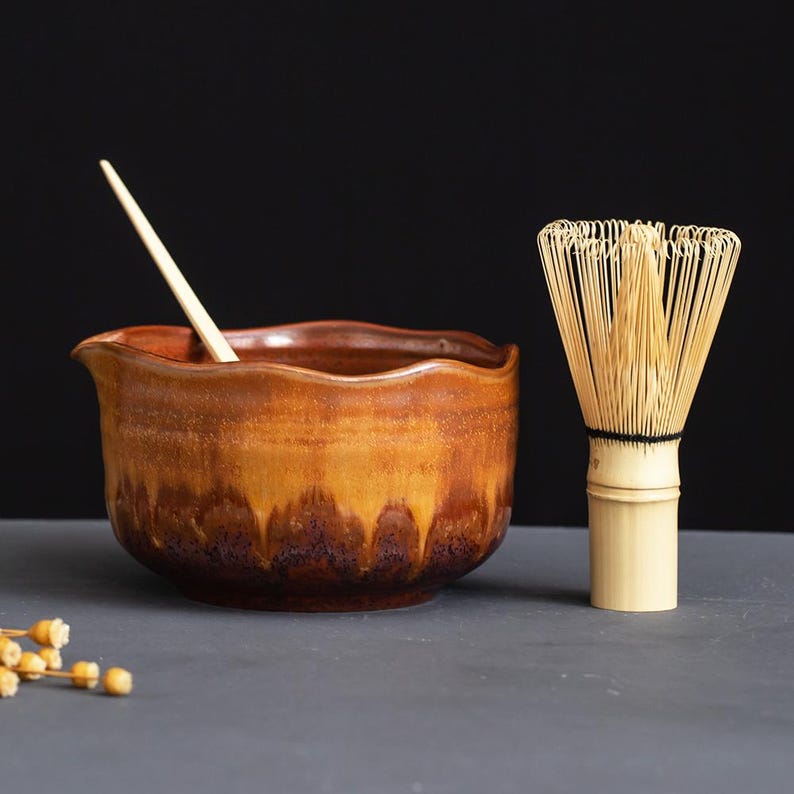 May include: A ceramic matcha bowl with a wavy rim and a brown and amber drip glaze, accompanied by a light-colored bamboo whisk and a thin wooden stirring stick. The bowl features a deep, mottled base that transitions into a lighter golden hue.
