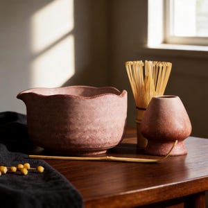 Könnte beinhalten: Eine rosa Matcha-Schale aus Keramik mit gewelltem Rand und Ausguss, ein Bambusbesen, ein Bambuslöffel und ein rosa Keramikhalter für den Besen. Das Set zeichnet sich durch eine texturierte, gesprenkelte Oberfläche der Keramikteile aus.