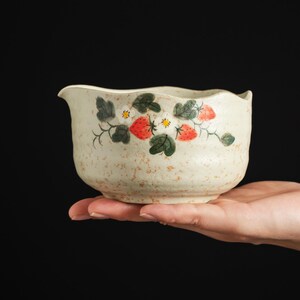 May include: A cream-colored ceramic bowl with a speckled texture and a hand-painted design featuring red strawberries, green leaves, and small white flowers. The bowl has a slightly wavy rim and a small pouring spout on one side of the vessel.