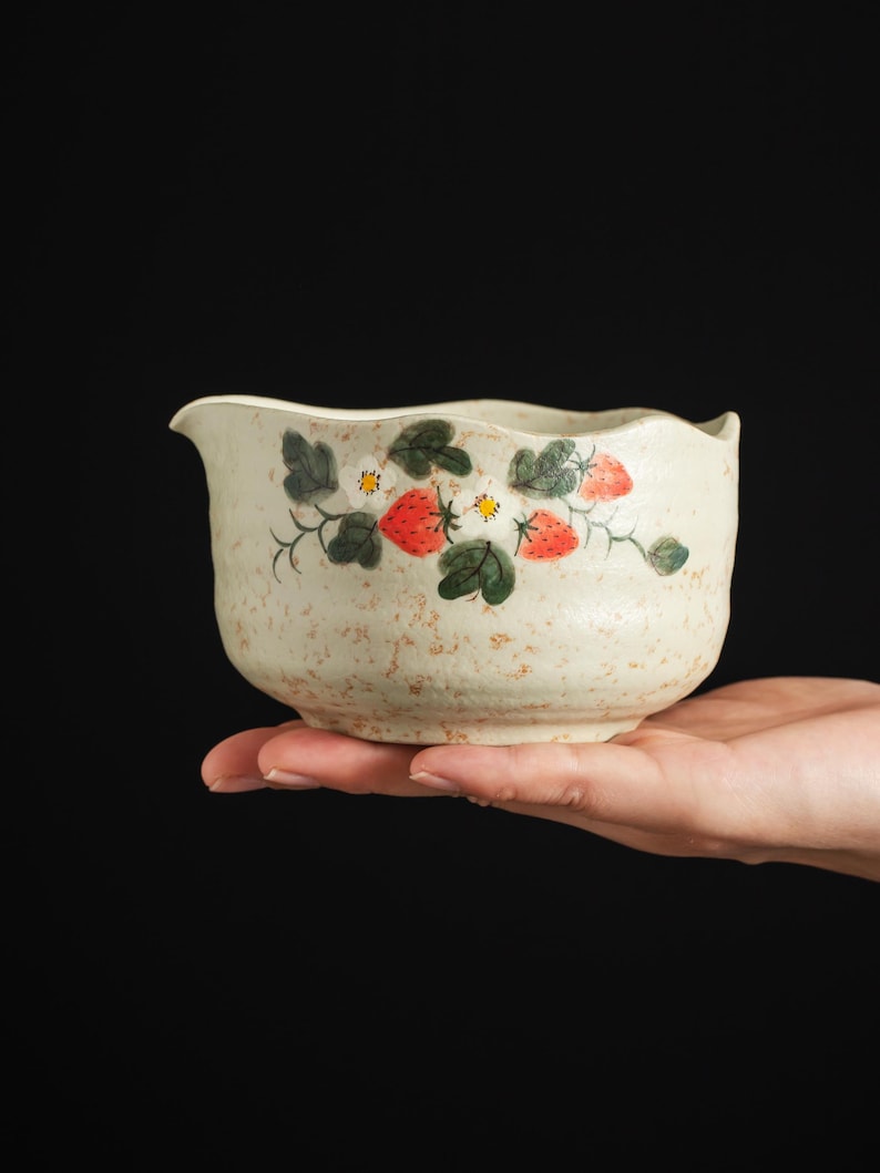 May include: A cream-colored ceramic bowl with a speckled texture and a hand-painted design featuring red strawberries, green leaves, and small white flowers. The bowl has a slightly wavy rim and a small pouring spout on one side of the vessel.