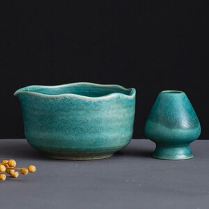 May include: A teal ceramic tea bowl with a wavy rim and a small, matching teal whisk holder. The bowl has a spout and a textured surface. The whisk holder is a small, rounded shape. The items are on a gray surface.