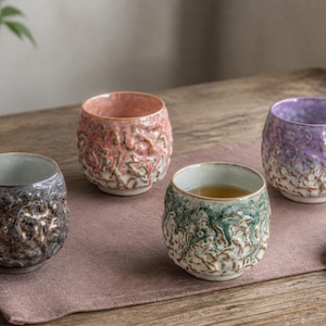 May include: Four ceramic tea cups with a textured, organic exterior surface. The cups feature distinct color glazes in dark grey, soft pink, light green, and lavender. Each cup has a smooth interior and a rounded shape, with one cup containing tea.