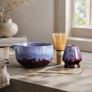 Puede incluir: Un cuenco de cerámica para matcha con esmalte de goteo azul y rojo oscuro, un batidor de bambú, un soporte de cerámica a juego y una cuchara de té de bambú. El cuenco tiene un pequeño pico vertedor en un lado y un degradado de colores.