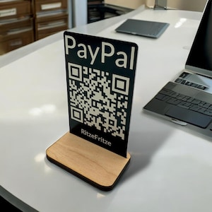 Könnte beinhalten: Ein schwarz-weißes PayPal QR-Code-Schild mit Holzsockel. Das Schild zeigt das PayPal-Logo und einen scannbaren QR-Code. Der Text "RitzeFritze" ist am unteren Rand des Schildes aufgedruckt. Das Schild steht auf einer weißen Oberfläche.