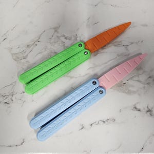 May include: Two plastic butterfly knives, one green and one blue, with orange and pink blades. The knives are on a white marble surface.