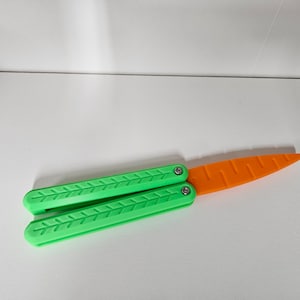 May include: A green and orange plastic toy knife shaped like a butterfly knife. The blade is orange and the handle is green.