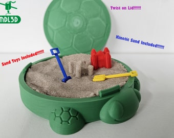Puede incluir: Caja de arena verde en forma de tortuga con una tapa que se abre para revelar un área de juego de arena. La caja de arena incluye un castillo rojo, un molde para castillo de arena, una pala azul y un rastrillo amarillo. La tapa tiene un diseño de balón de fútbol. El texto "Twist on Lid!!!" está escrito en morado encima de la caja de arena y el texto "Kinetic Sand Included!!!!" está escrito en morado debajo de la caja de arena. El texto "Sand Toys Included!!!!" está escrito en rojo a la izquierda de la caja de arena.
